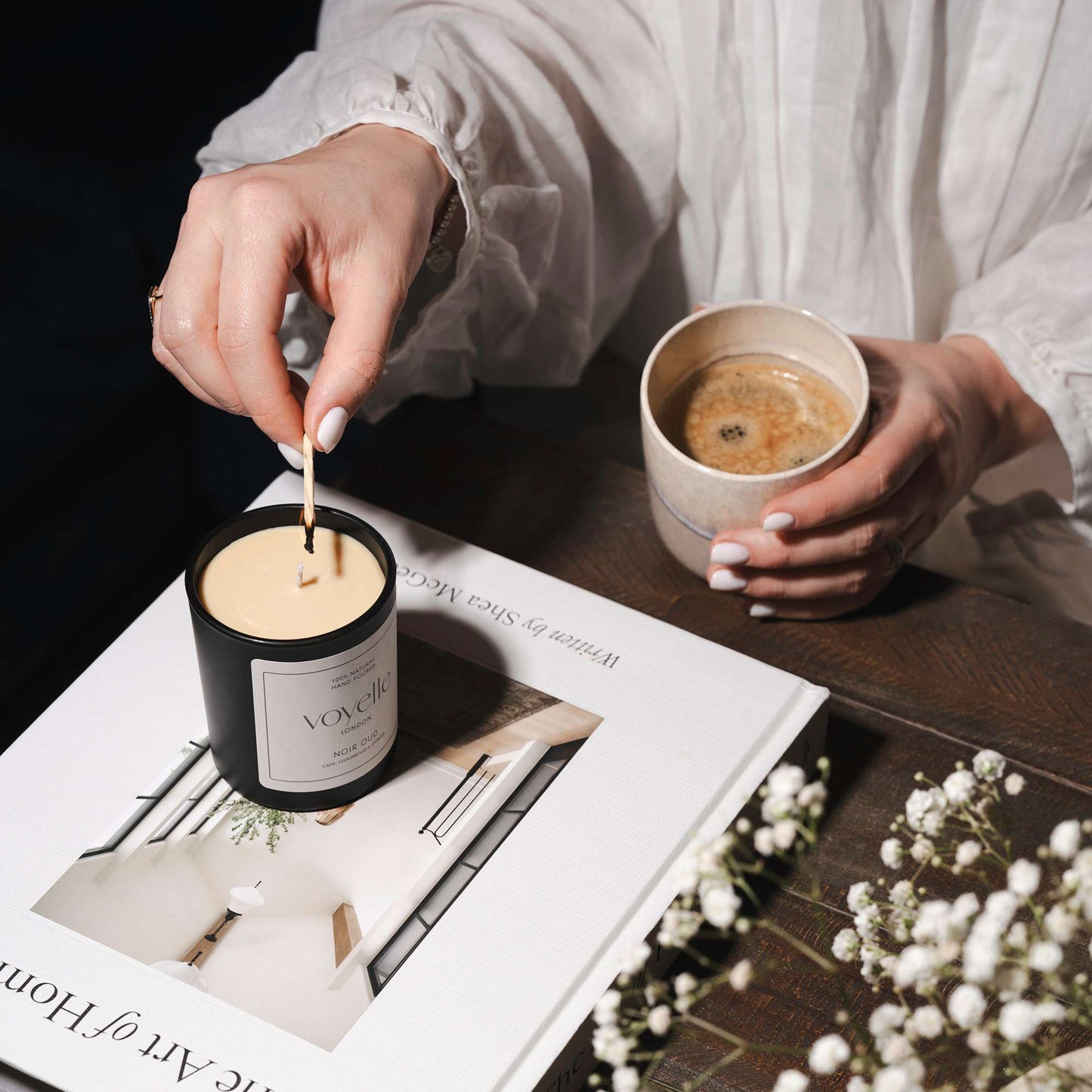 Person lighting a black Voyelle candle with a match while holding a cup of coffee, placed on a coffee table with a book and white flowers