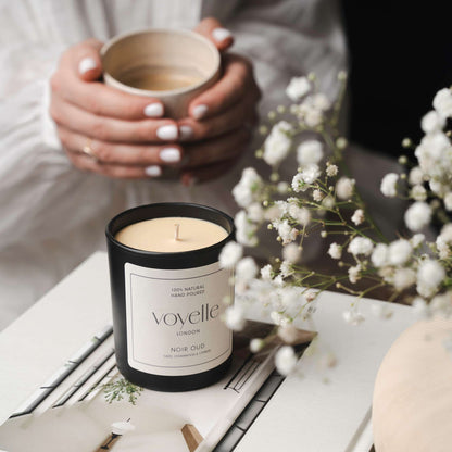 Black Voyelle candle placed on a book next to delicate white flowers, with a person holding a coffee cup in the background