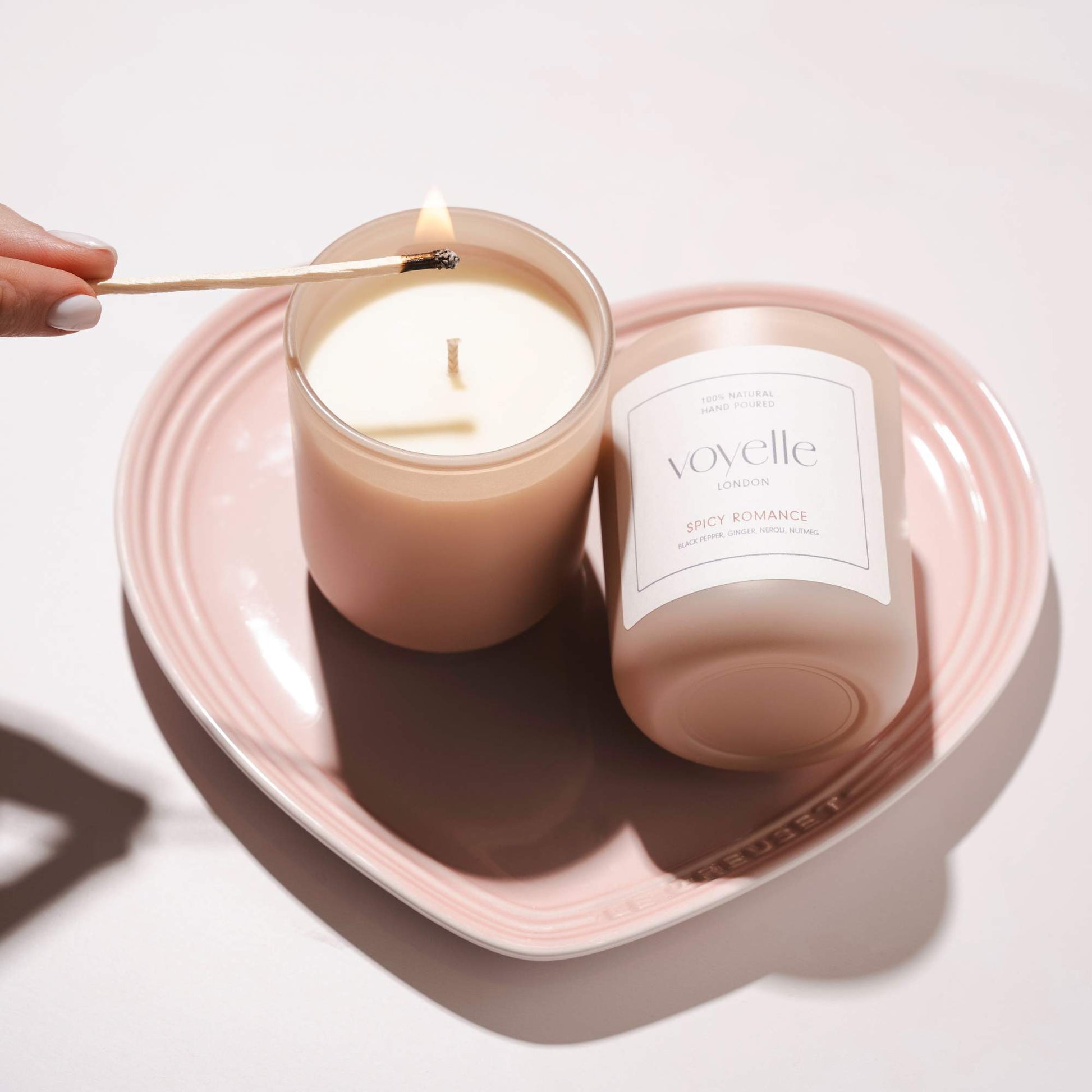 Hand lighting a Voyelle Spicy Romance candle on a pink heart-shaped plate