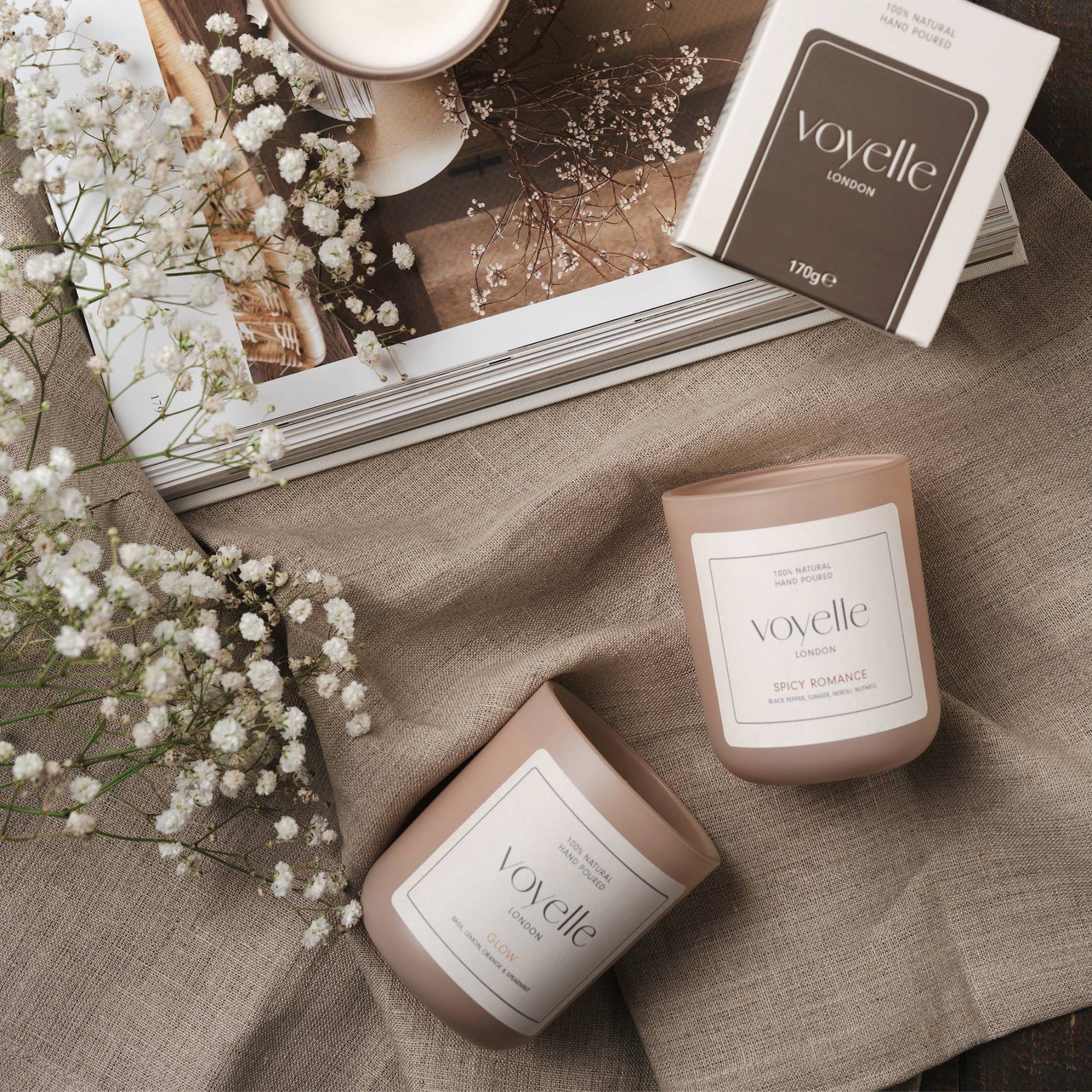 Two Voyelle candles on fabric with a book and flowers in the background