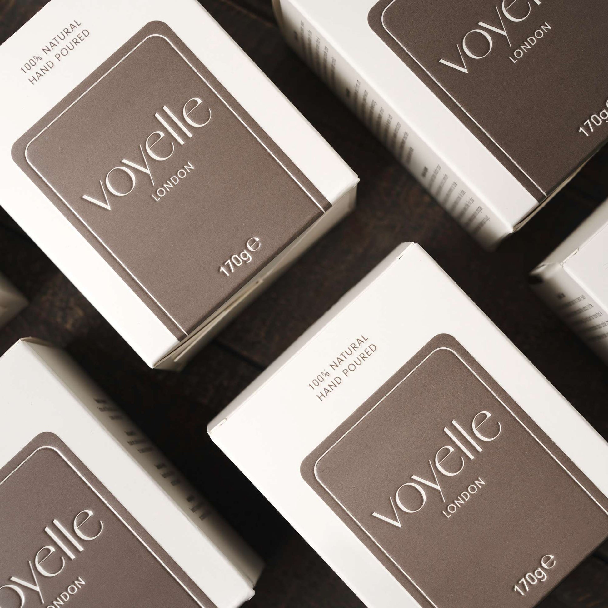 Close-up of multiple Voyelle London candle boxes in brown and white packaging on a wooden surface