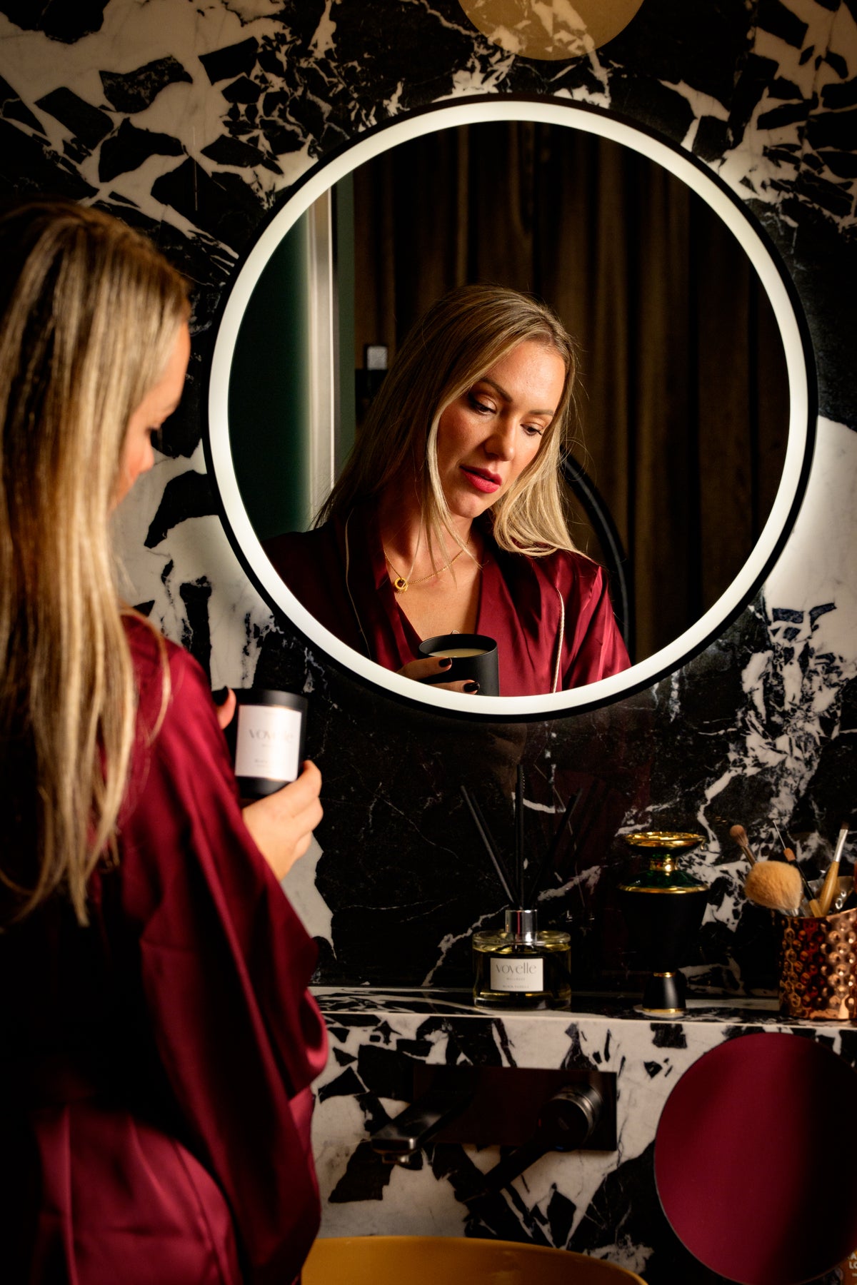 Karolina in a burgundy silk robe holds a Voyelle candle at a round lit mirror on a marble vanity