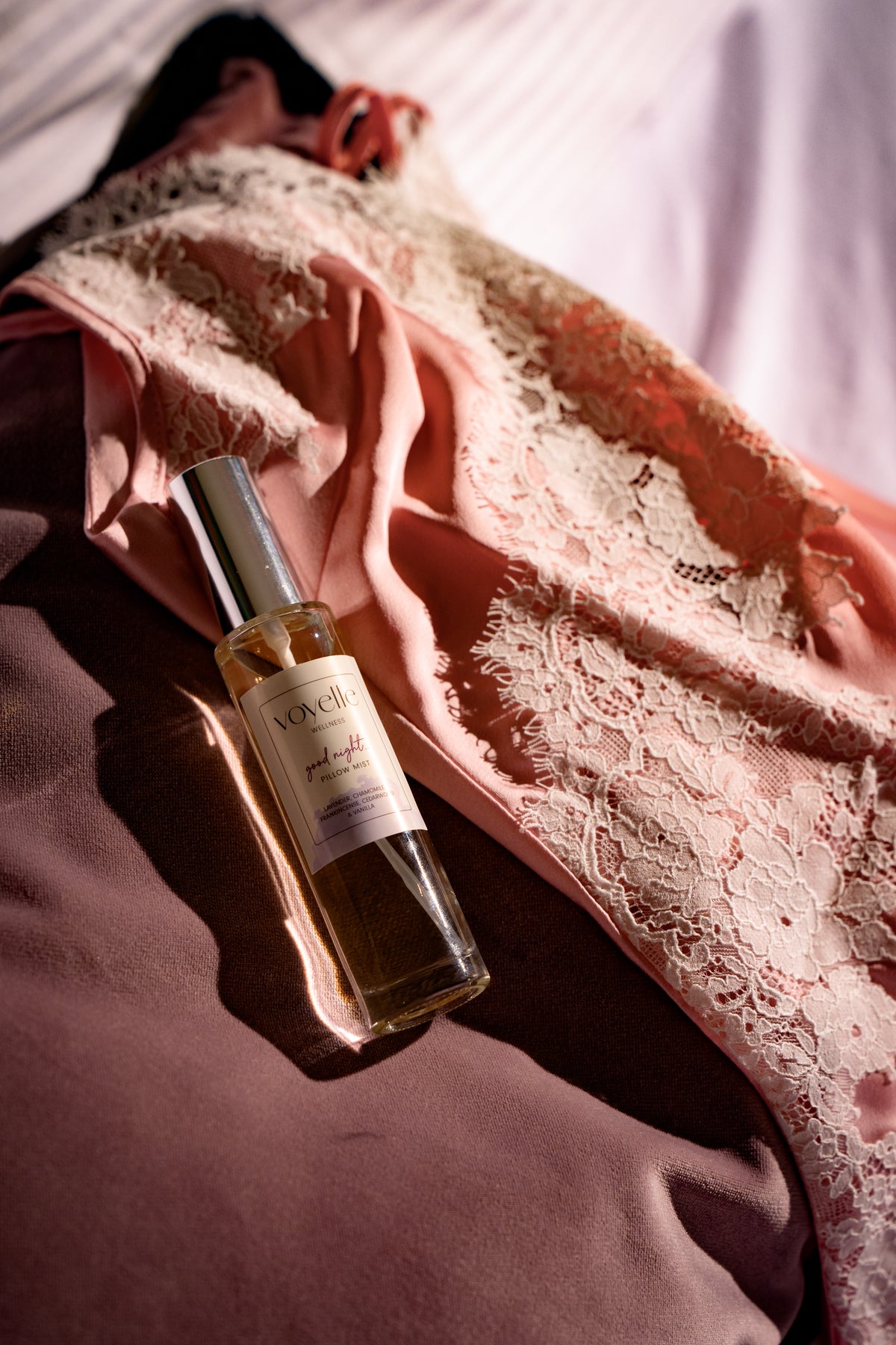 A clear glass bottle of Voyelle Pillow Mist with a silver cap, resting on draped pink satin and lace fabric. For better sleep