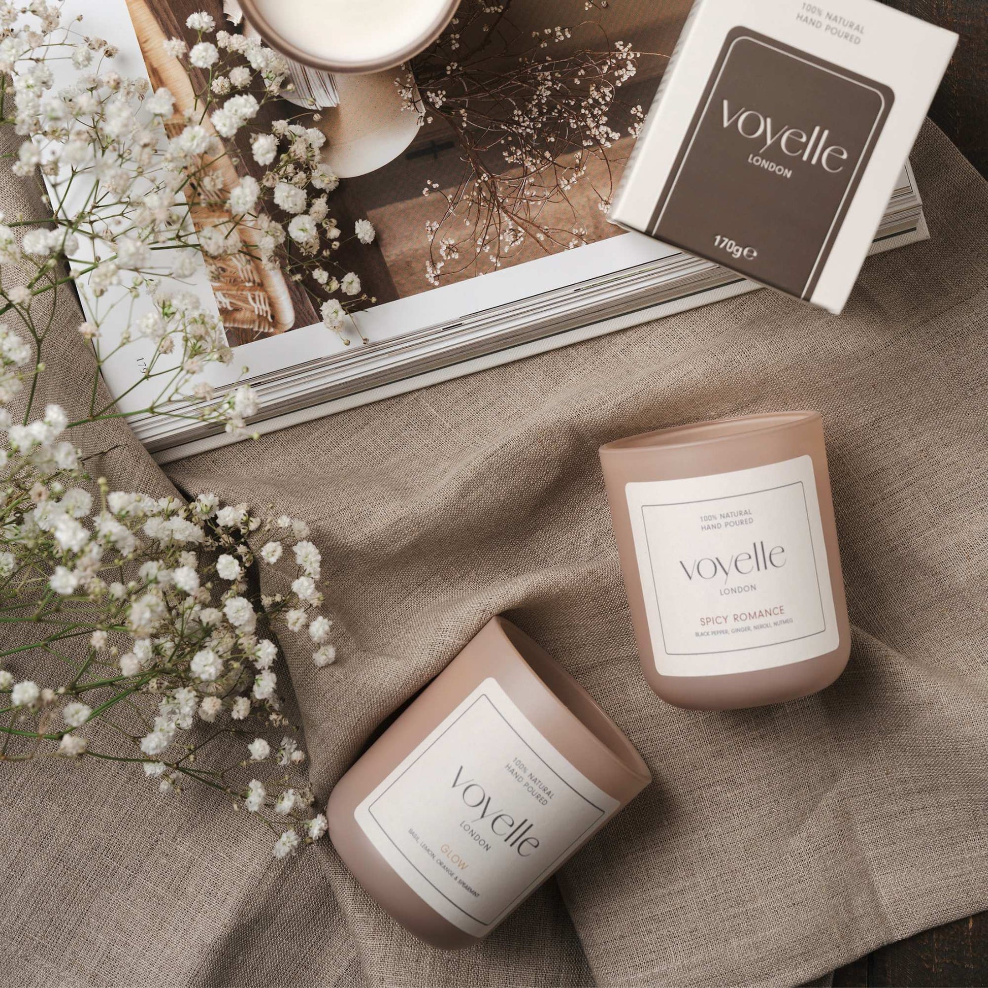 Two Voyelle candles – Glow and Spicy Romance – styled with linen, flowers, and an open magazine