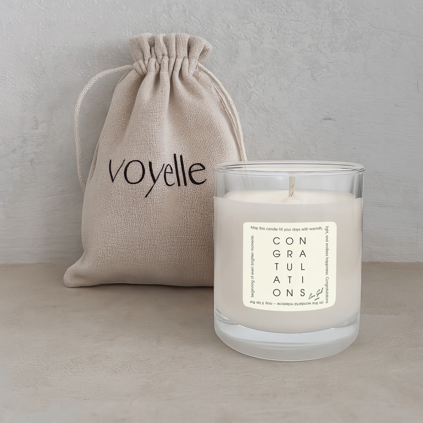 Candle with 'Congratulations' label and beige drawstring bag labeled 'voyelle' on a light background