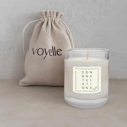 Candle with 'Congratulations' label and beige drawstring bag labeled 'voyelle' on a light background