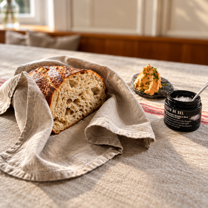 A rustic loaf of sourdough bread on a beige linen cloth, with a small jar of salt and a spoonful of orange spread in the background on a table in a warm indoor setting.
