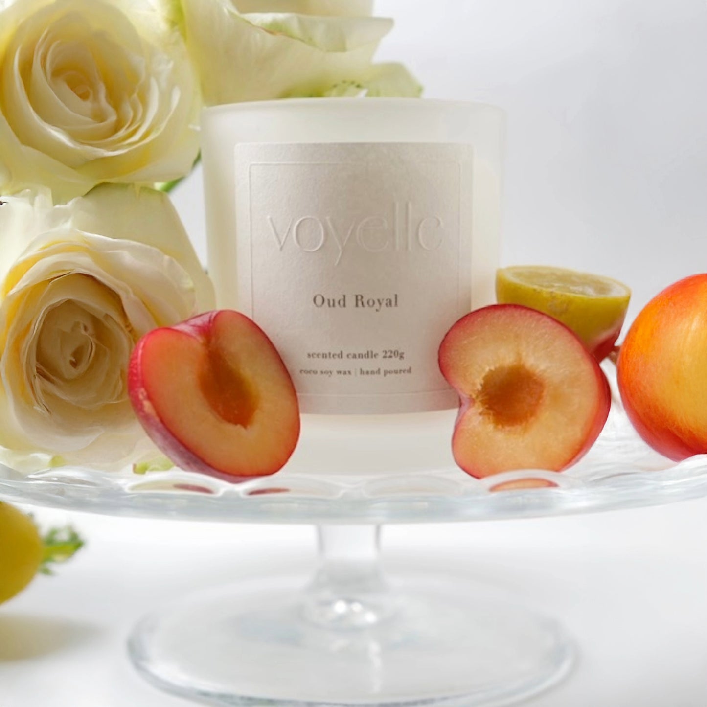 Voyelle Oud Royal candle with sliced plums, lemon and white roses on a glass stand