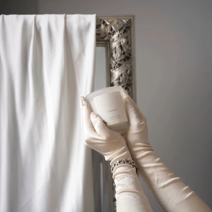 Gloved hands holding Voyelle Oud Royal candle in front of ornate mirror and white curtain