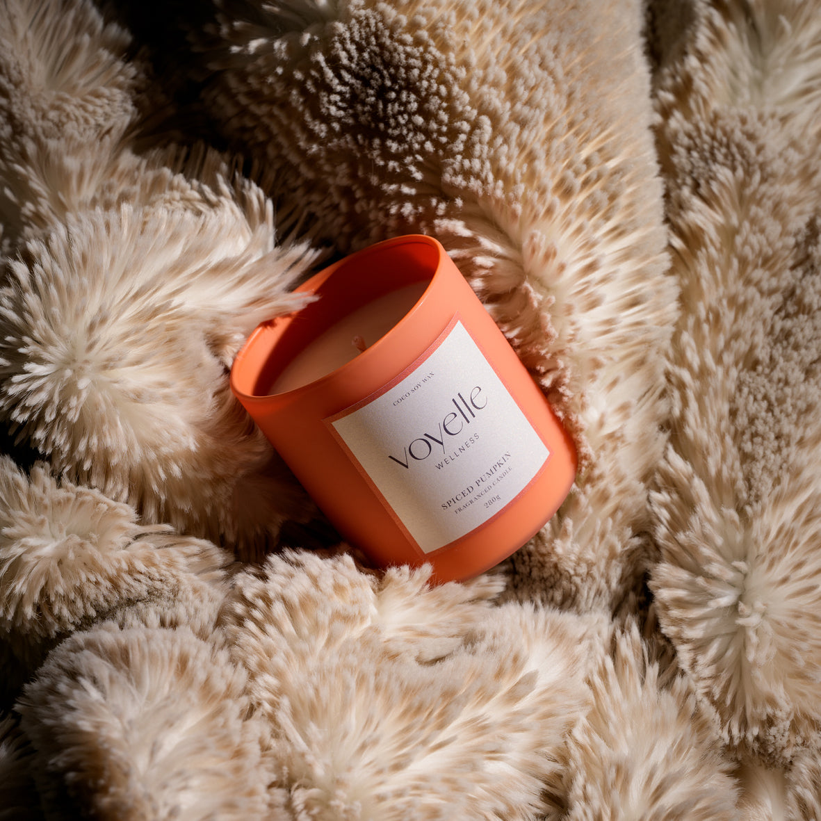 An orange cylindrical candle jar with a white label reading 'Voyelle' and 'Sapin de Coco' resting on a fluffy cream faux-fur surface.