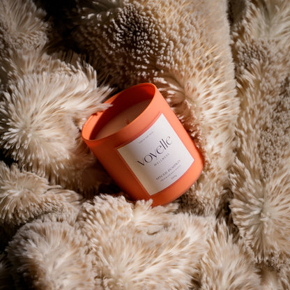An orange cylindrical candle jar with a white label reading 'Voyelle' and 'Sapin de Coco' resting on a fluffy cream faux-fur surface.