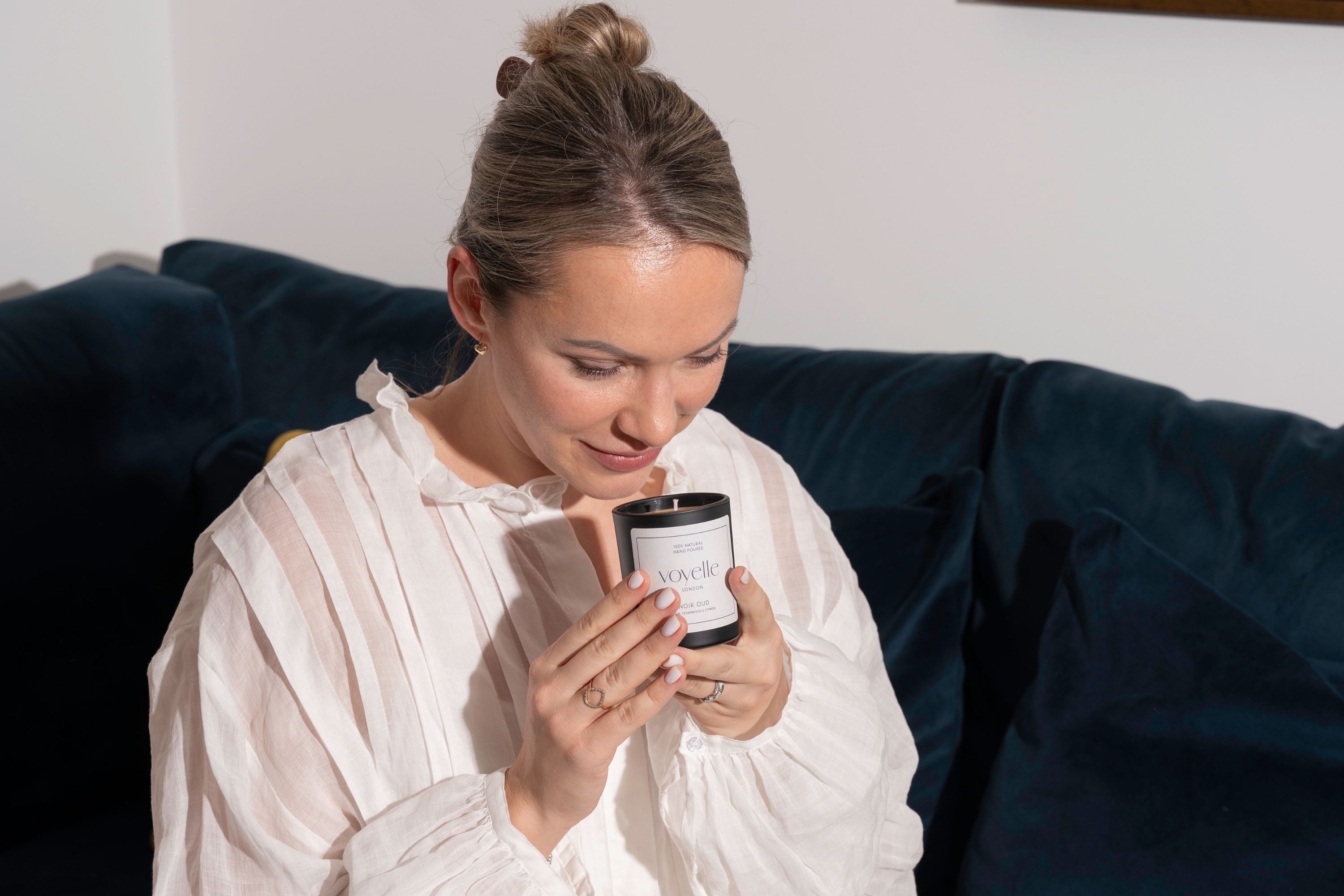 Woman smelling Voyelle scented candle while relaxing on a navy blue sofa – luxury aromatherapy candle experience